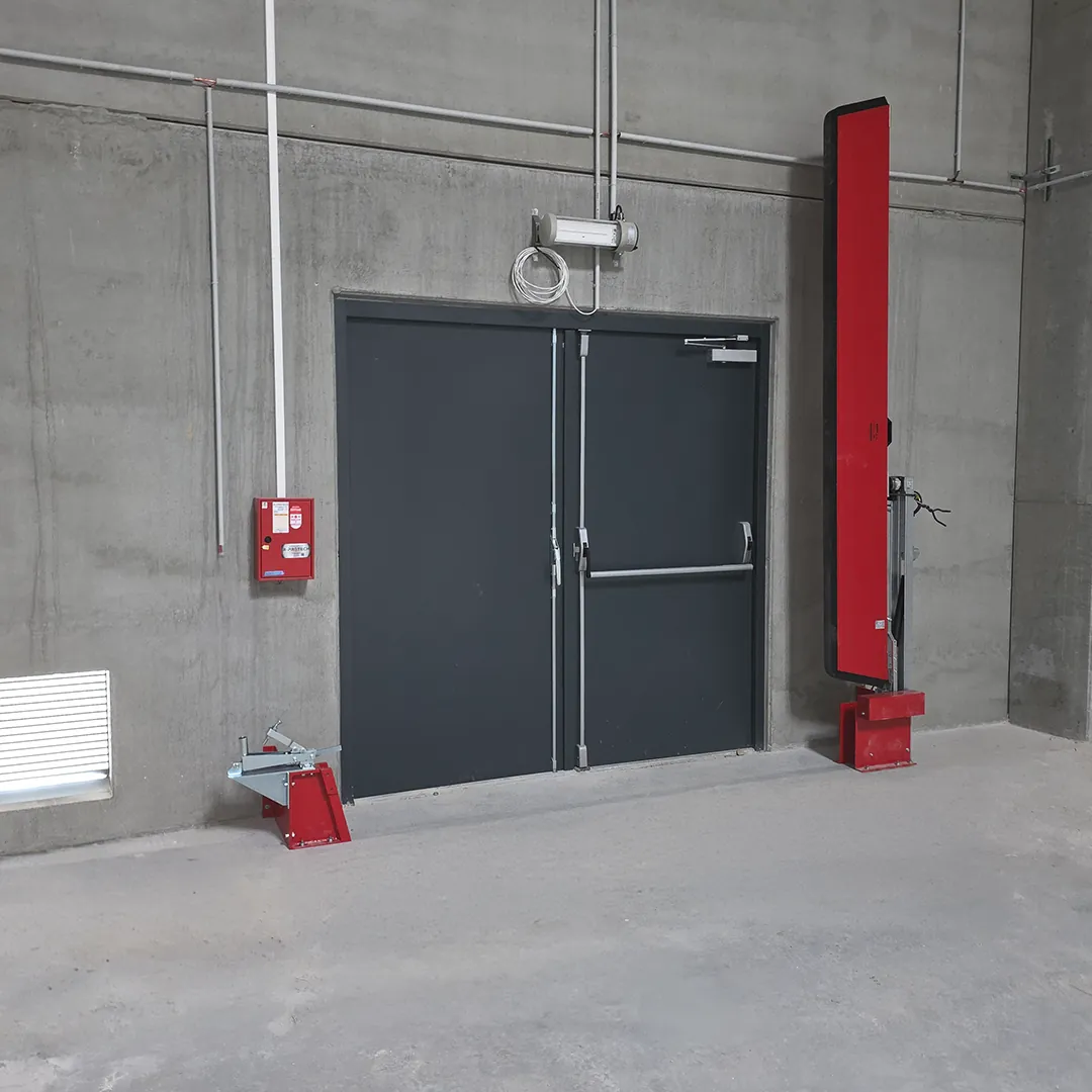Barrière de rétention automatique rouge relevée à côté d'une porte double, avec son support au sol de l'autre côté