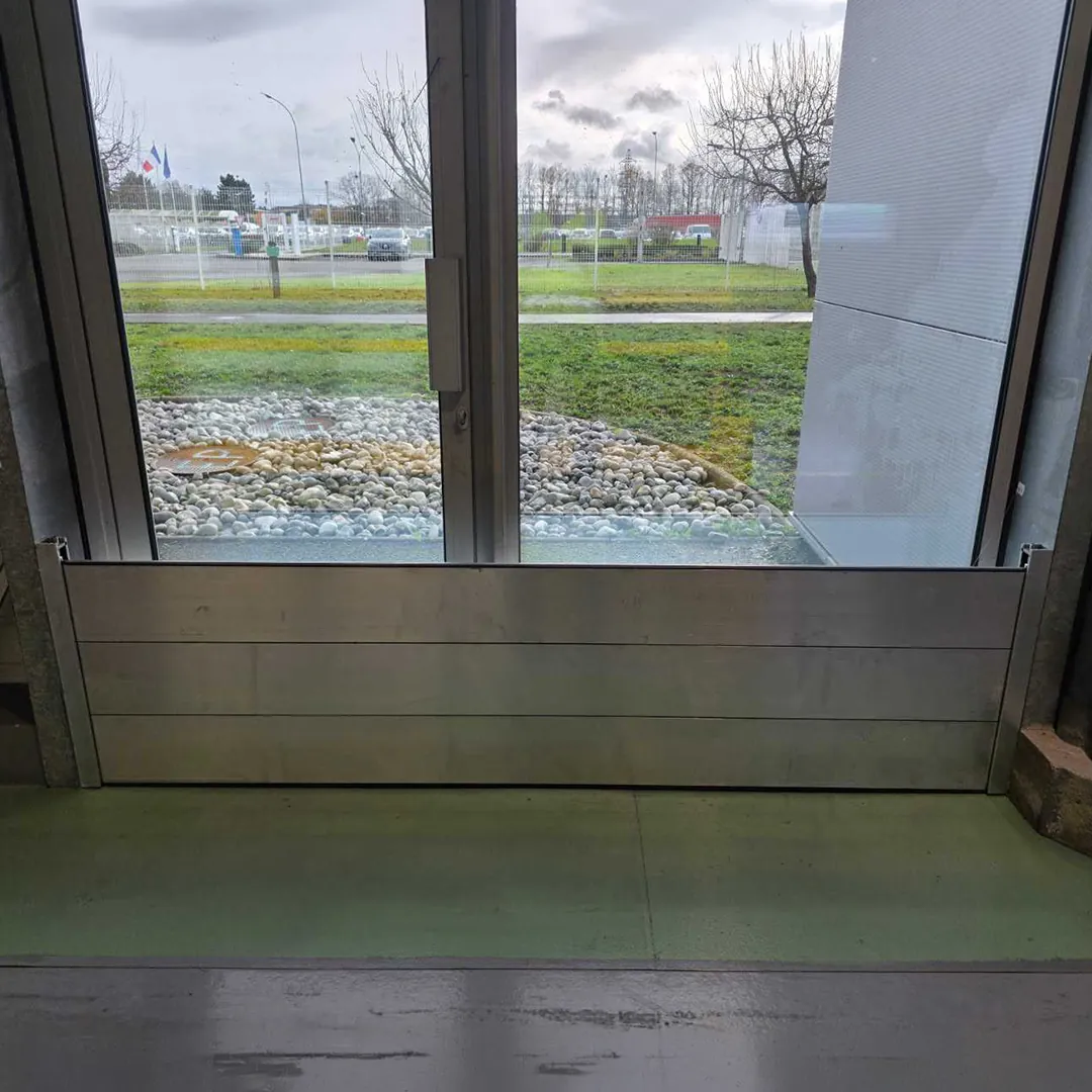 Batardeau aluminium à lames devant une baie vitrée, vu depuis l'intérieur du bâtiment